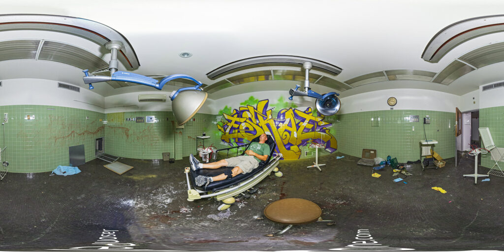 A 360-degree panoramic image inside an abandoned hospital showing the photographer on an operating table with graffiti behind the table.
